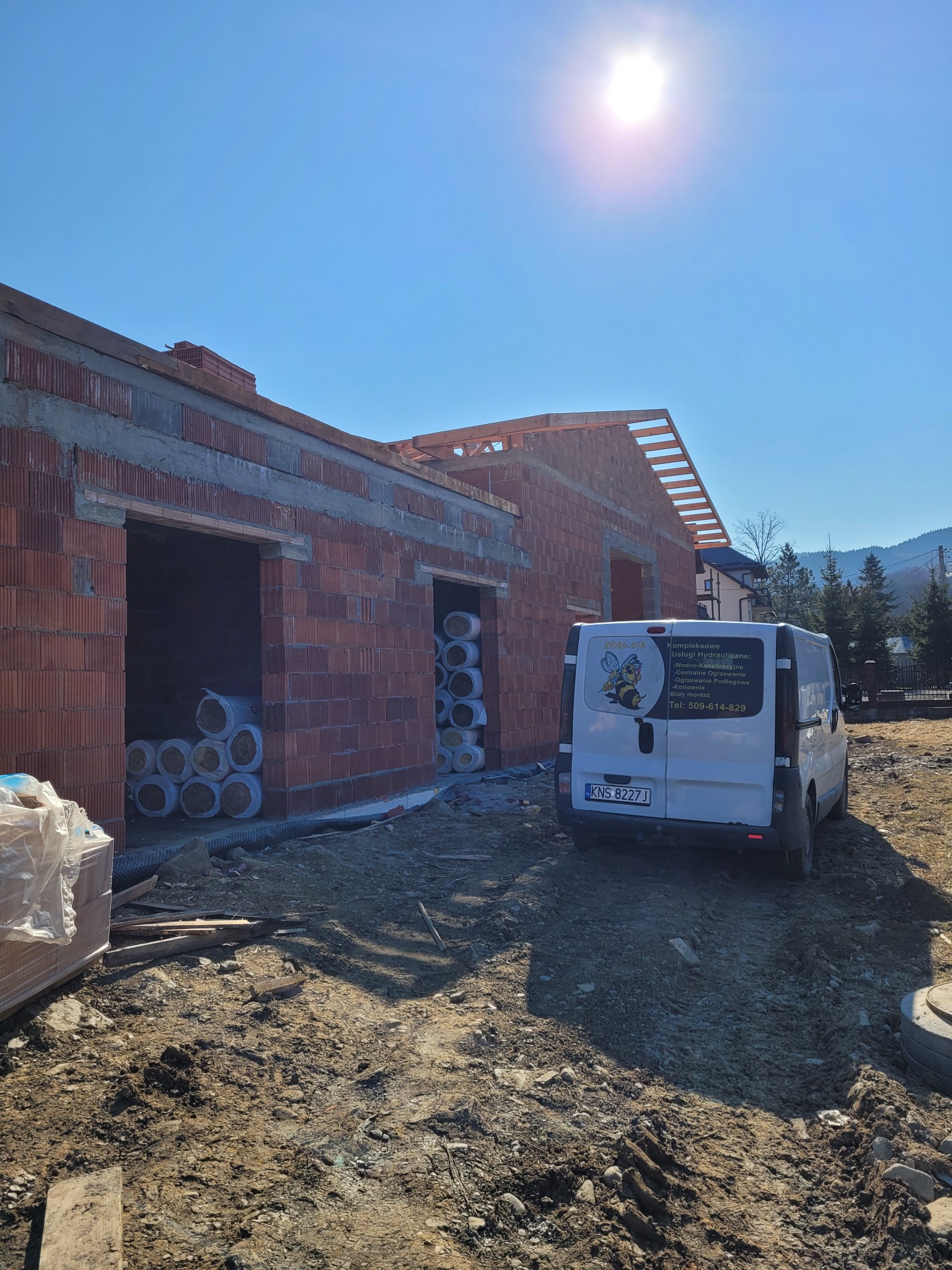 Budowa domu z czerwonej cegły w trakcie realizacji, widoczny samochód dostawczy z reklamą usług instalacyjnych na tle górskiego krajobrazu.
