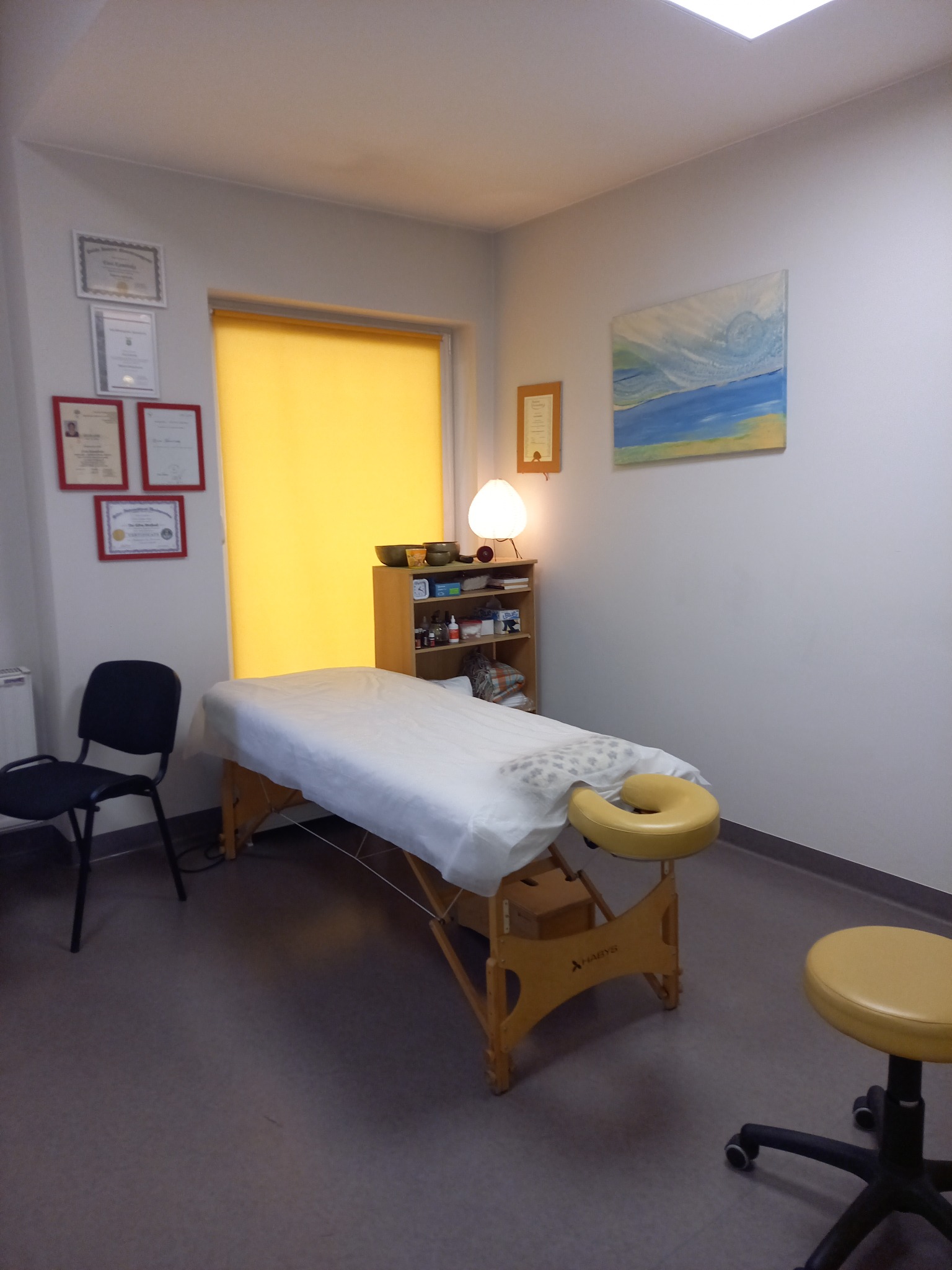 Jasne wnętrze gabinetu z kozetką do masażu, krzesłem i półkami. Na ścianie wiszą dyplomy i obraz. Okno zasłonięte żółtą roletą.