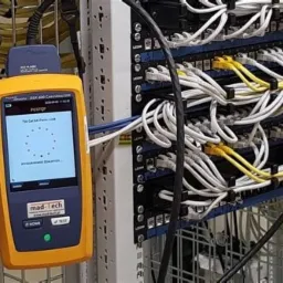 Miernik Fluke DSX-602 CableAnalyzer podczas testowania okablowania strukturalnego w szafie rack, widoczne patch panele i kable zakończone wtykami RJ45.