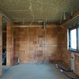 Wnętrze w stanie surowym: instalacja elektryczna w trakcie montażu, widoczne przewody rozprowadzone po ścianach z cegły, rury wentylacyjne pod sufitem, okno z parapetem, niebieskie rury na podłodze.