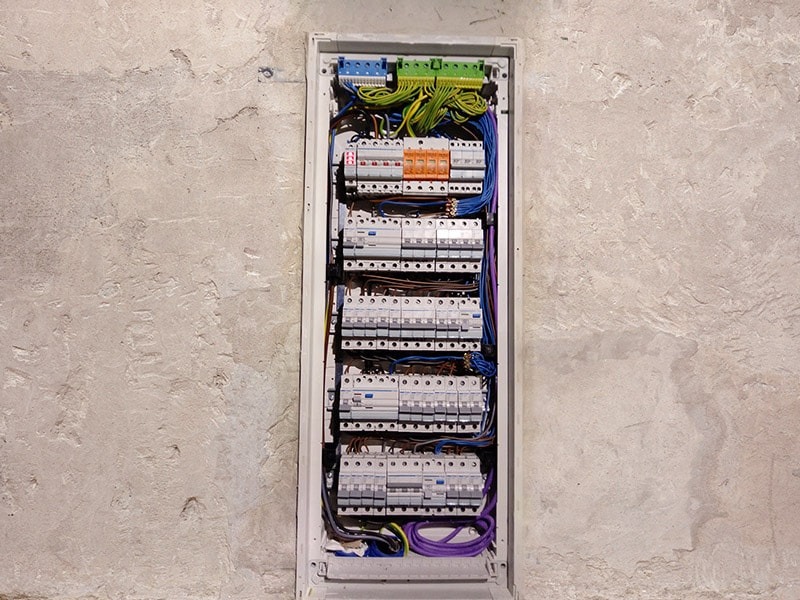 Rozdzielnica elektryczna z zainstalowanymi bezpiecznikami i okablowaniem, widoczna na tle surowej ściany z betonu architektonicznego.