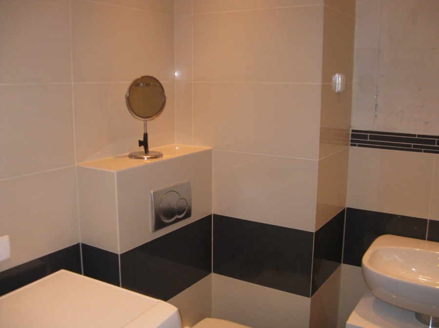 Wykończona łazienka z beżowo-czarnymi płytkami, wiszącą umywalką i okrągłym lustrem na blacie zabudowy stelaża WC. Widoczny chromowany przycisk spłuczki.