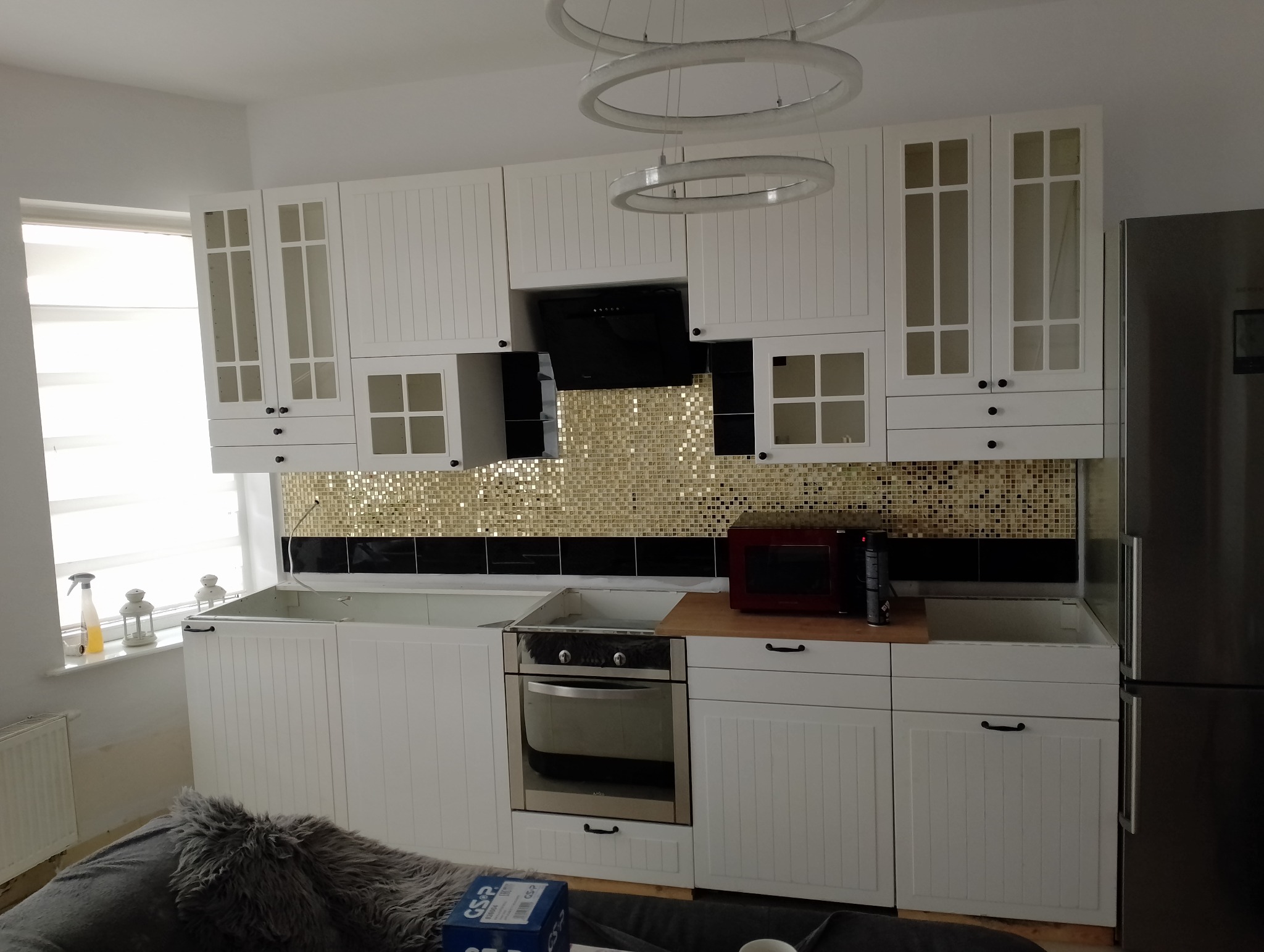 Białe szafki kuchenne z czarnymi uchwytami, mozaika w odcieniach złota i czerni nad blatem, brak zlewu i baterii, widoczny piekarnik i okap, nowoczesne oświetlenie sufitowe.