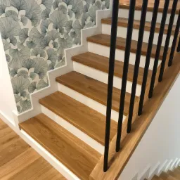 Drewniane schody z czarną balustradą i białymi podstopnicami, ściana z tapetą w motywie liści miłorzębu, widok z dołu.