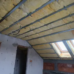 Poddasze w trakcie izolacji: wełna mineralna między metalowymi profilami sufitowymi, widoczne okno dachowe i fragment muru z pustaków.