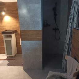 Damian Dworak DEKORACION - Nowoczesna łazienka z drewnopodobnymi płytkami na ścianie, szara kabina prysznicowa z czarną armaturą, umywalka z szafką w jasnym kolorze oraz krzesło z futrzanym siedziskiem