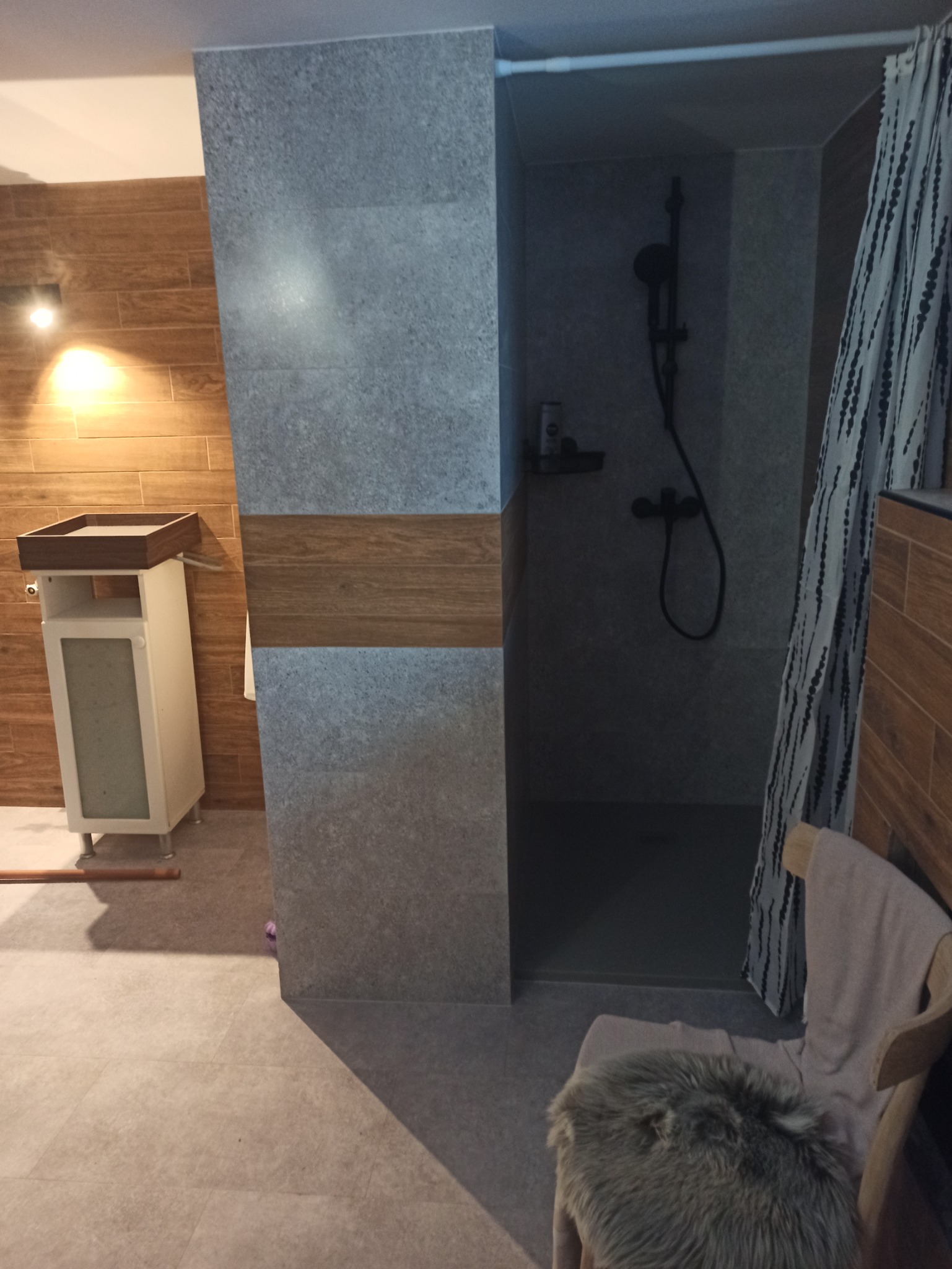 Nowoczesna łazienka z drewnopodobnymi płytkami na ścianie, szara kabina prysznicowa z czarną armaturą, umywalka z szafką w jasnym kolorze oraz krzesło z futrzanym siedziskiem