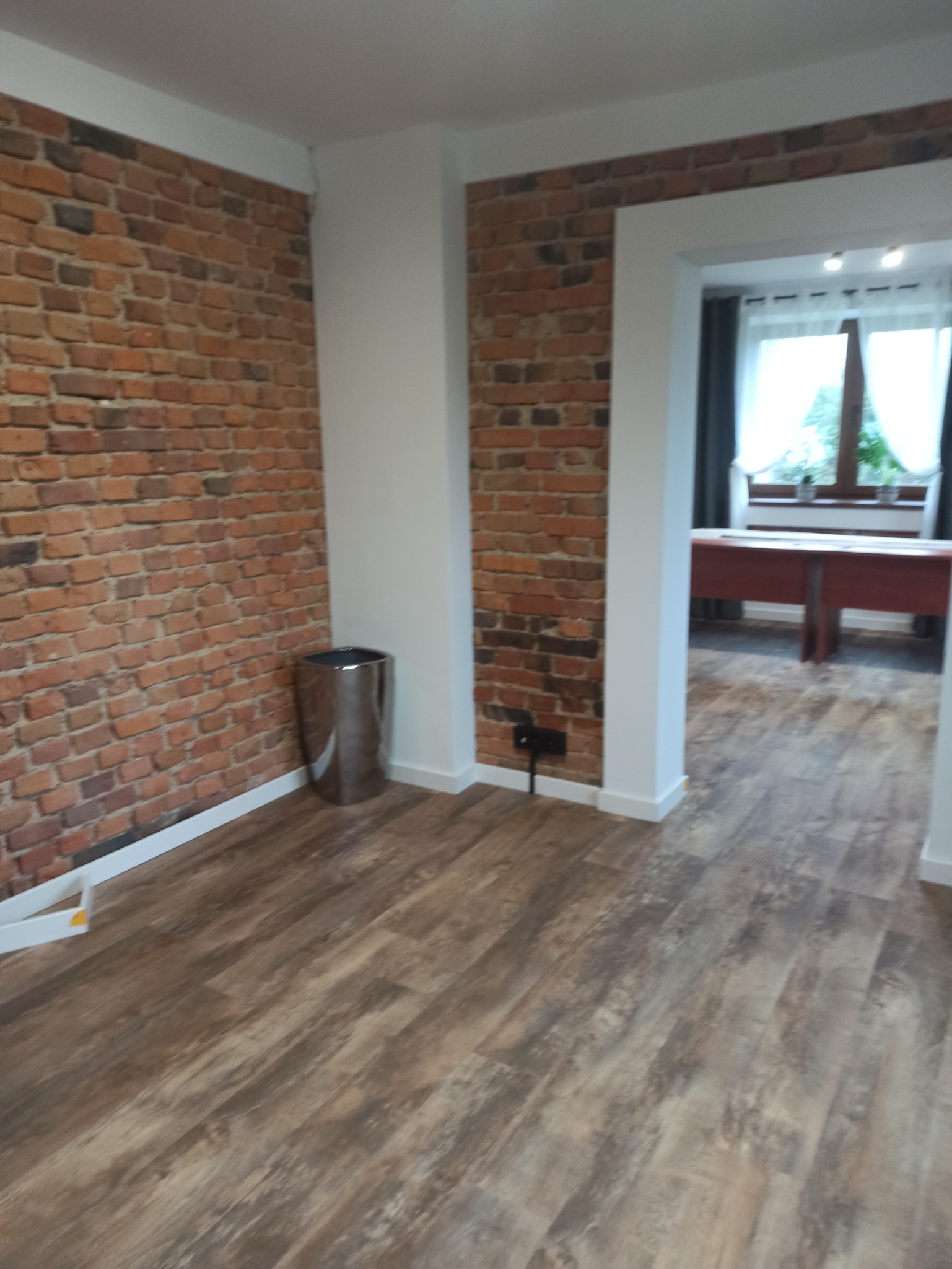 Pokój z drewnopodobną podłogą, ceglaną ścianą, srebrnym koszem na śmieci i widokiem na dalsze pomieszczenie z oknem.