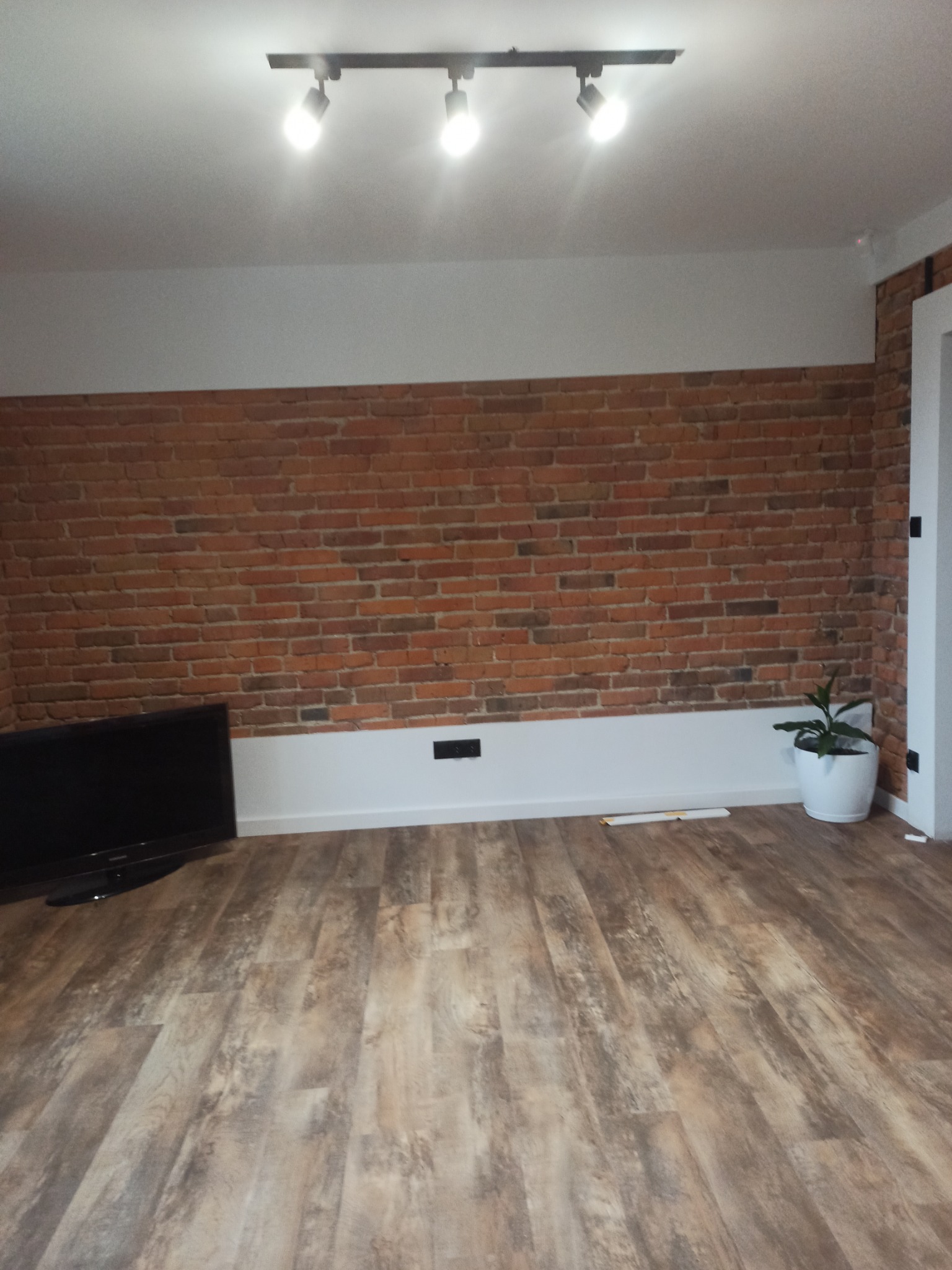 Salon po remoncie z drewnopodobną podłogą, ceglaną ścianą i nowoczesnym oświetleniem punktowym