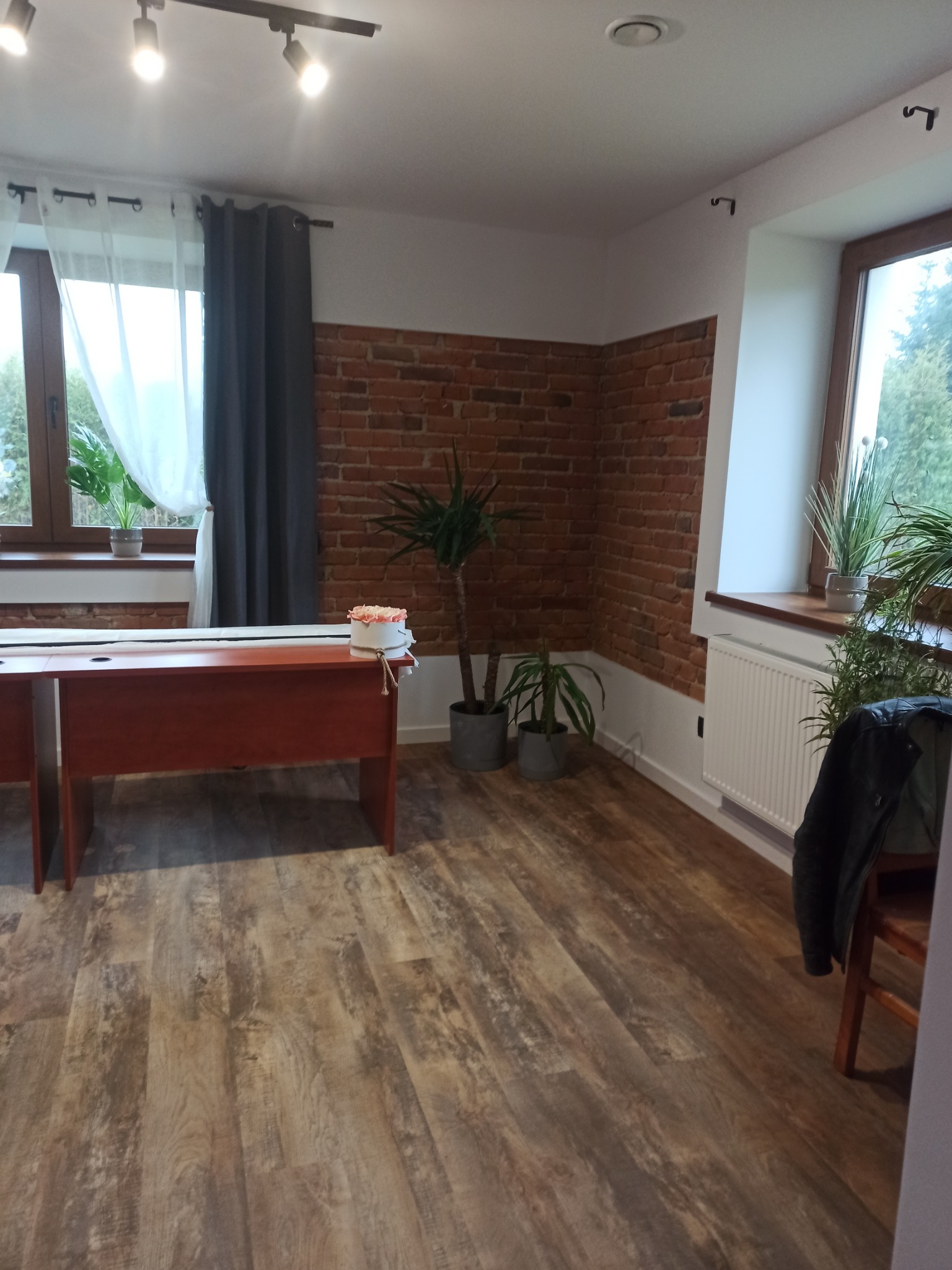 Pokój po remoncie z nową podłogą z paneli imitujących drewno, ściana z cegły, biurko z dekoracją kwiatową, okna z zasłonami i roślinami doniczkowymi.
