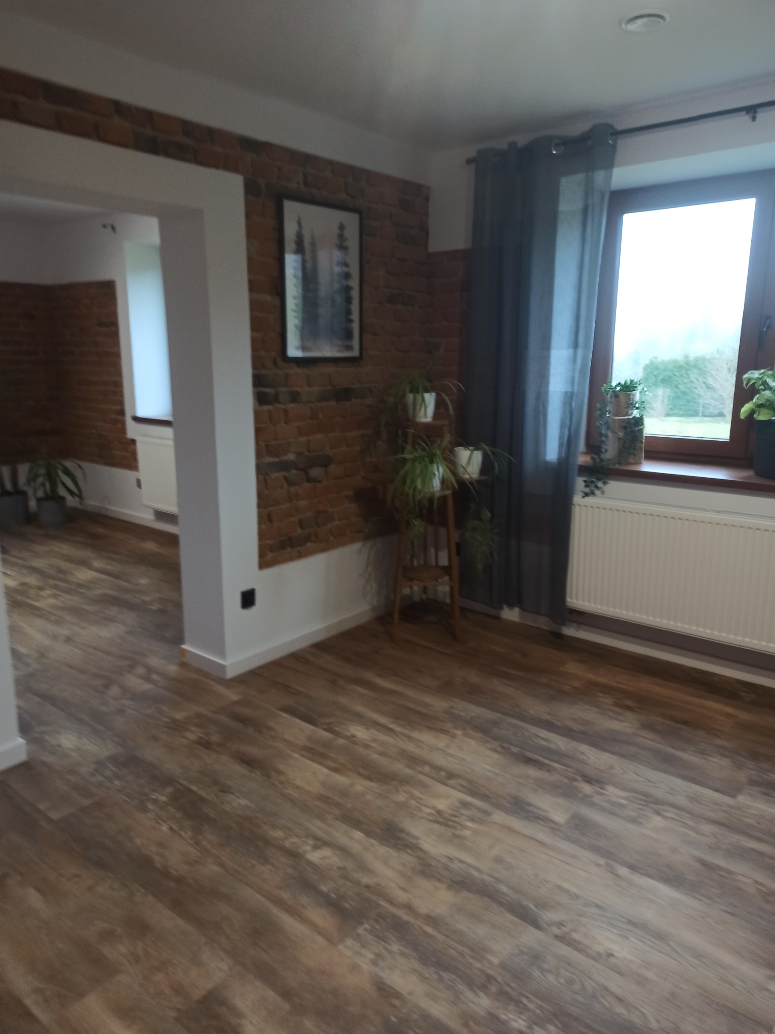 Pokój po remoncie z panelami podłogowymi w odcieniach brązu, ceglaną ścianą z obrazem i roślinami doniczkowymi na drewnianym kwietniku przy oknie z szarą zasłoną.