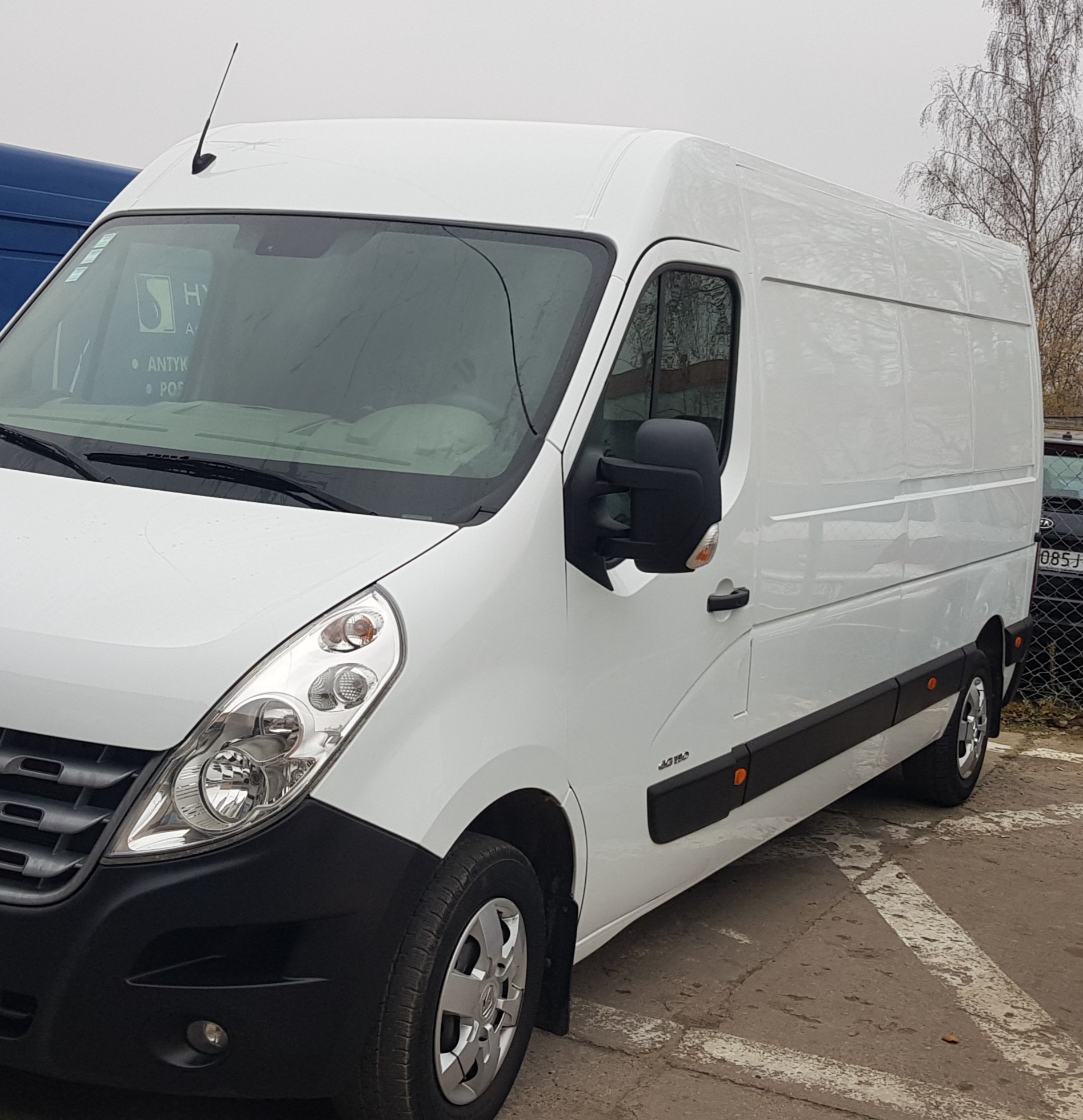 Biały samochód dostawczy Renault Master z anteną na dachu, zaparkowany na parkingu z widocznymi liniami, z logo firmy widocznym na przedniej szybie.