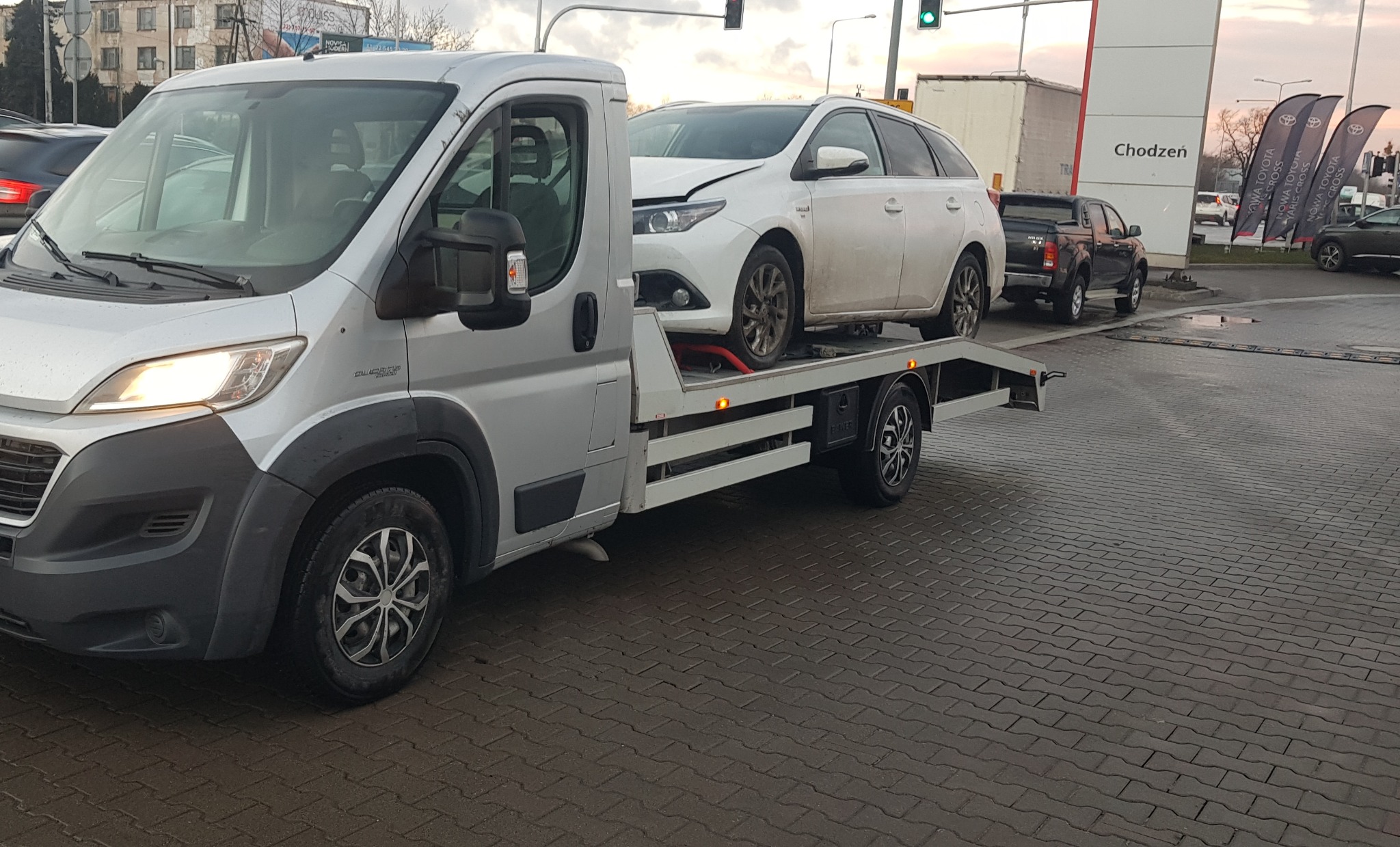 Srebrna laweta Fiat Ducato z uszkodzonym białym samochodem osobowym na platformie, stojąca na kostce brukowej przed salonem samochodowym Toyota Chodzeń.