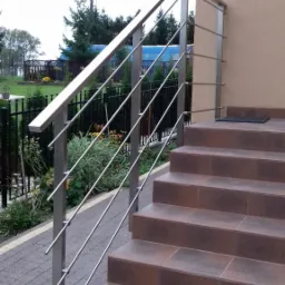 Zewnętrzne schody wejściowe wyłożone brązowymi płytkami, zabezpieczone nowoczesną, prostą balustradą ze stali nierdzewnej z poziomymi prętami, widok z dołu na tle zieleni.