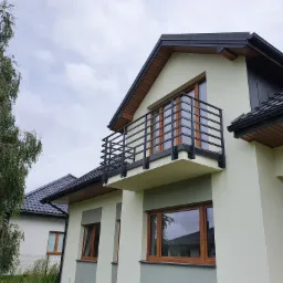 Nowoczesny dom z balkonem zabezpieczonym czarną, metalową balustradą o minimalistycznym designie, widok od dołu na tle pochmurnego nieba.