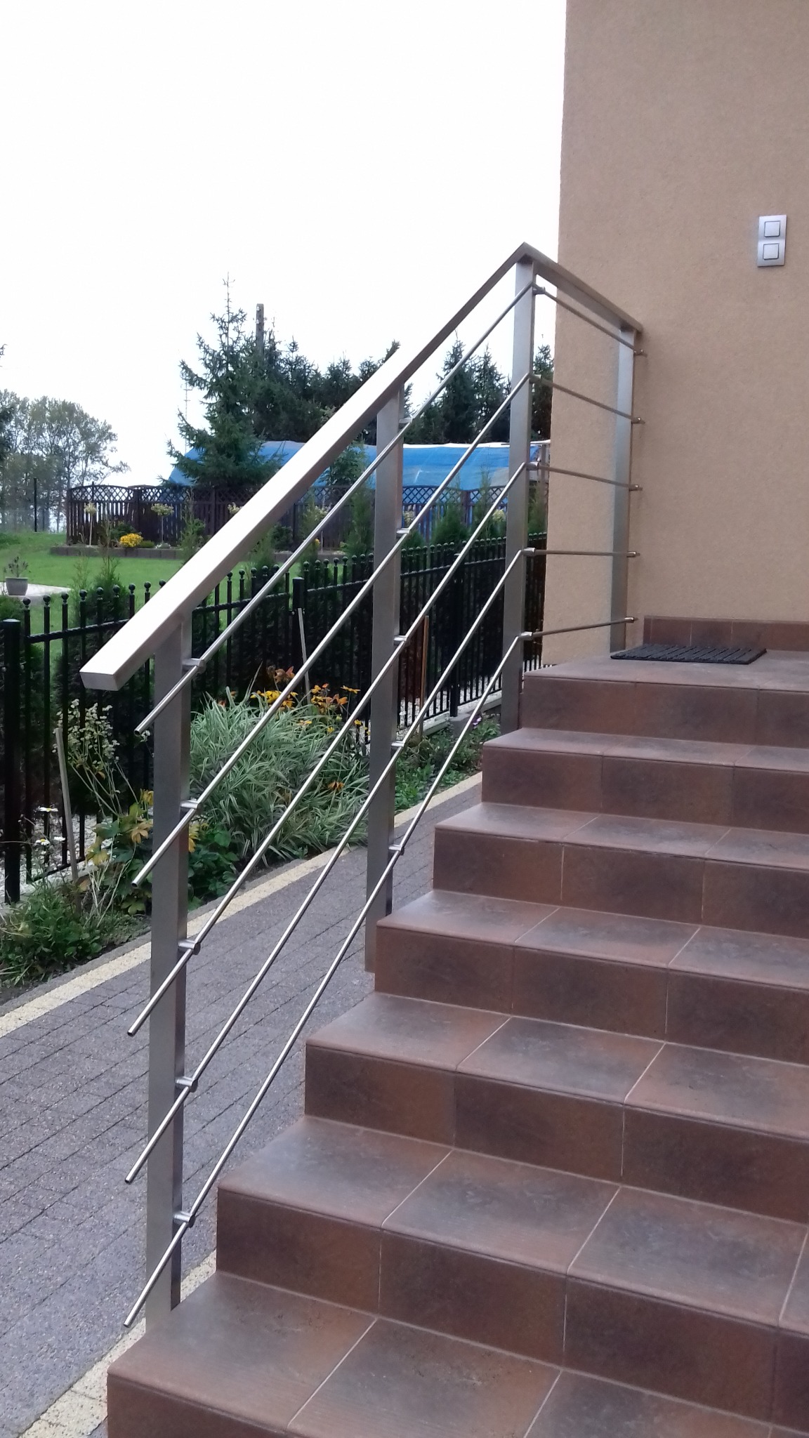 Zewnętrzne schody wejściowe wyłożone brązowymi płytkami, zabezpieczone nowoczesną, prostą balustradą ze stali nierdzewnej z poziomymi prętami, widok z dołu na tle zieleni.