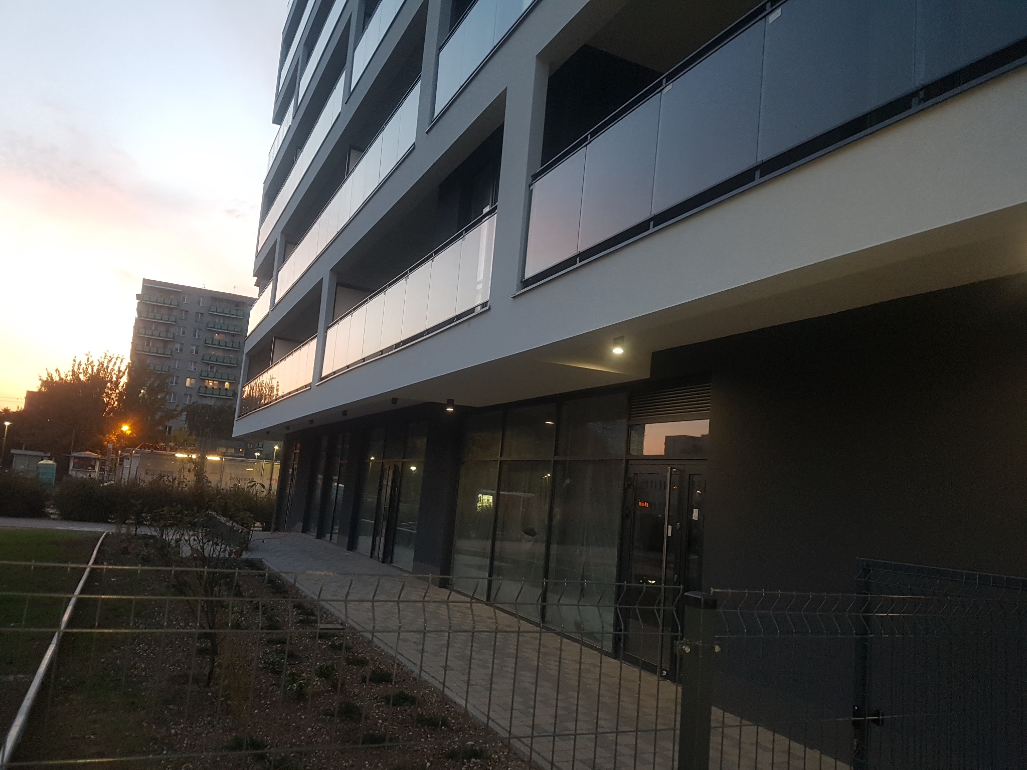 Nowoczesny budynek mieszkalny z balkonami zabezpieczonymi szkłem, widok z dołu na tle zachodzącego słońca.