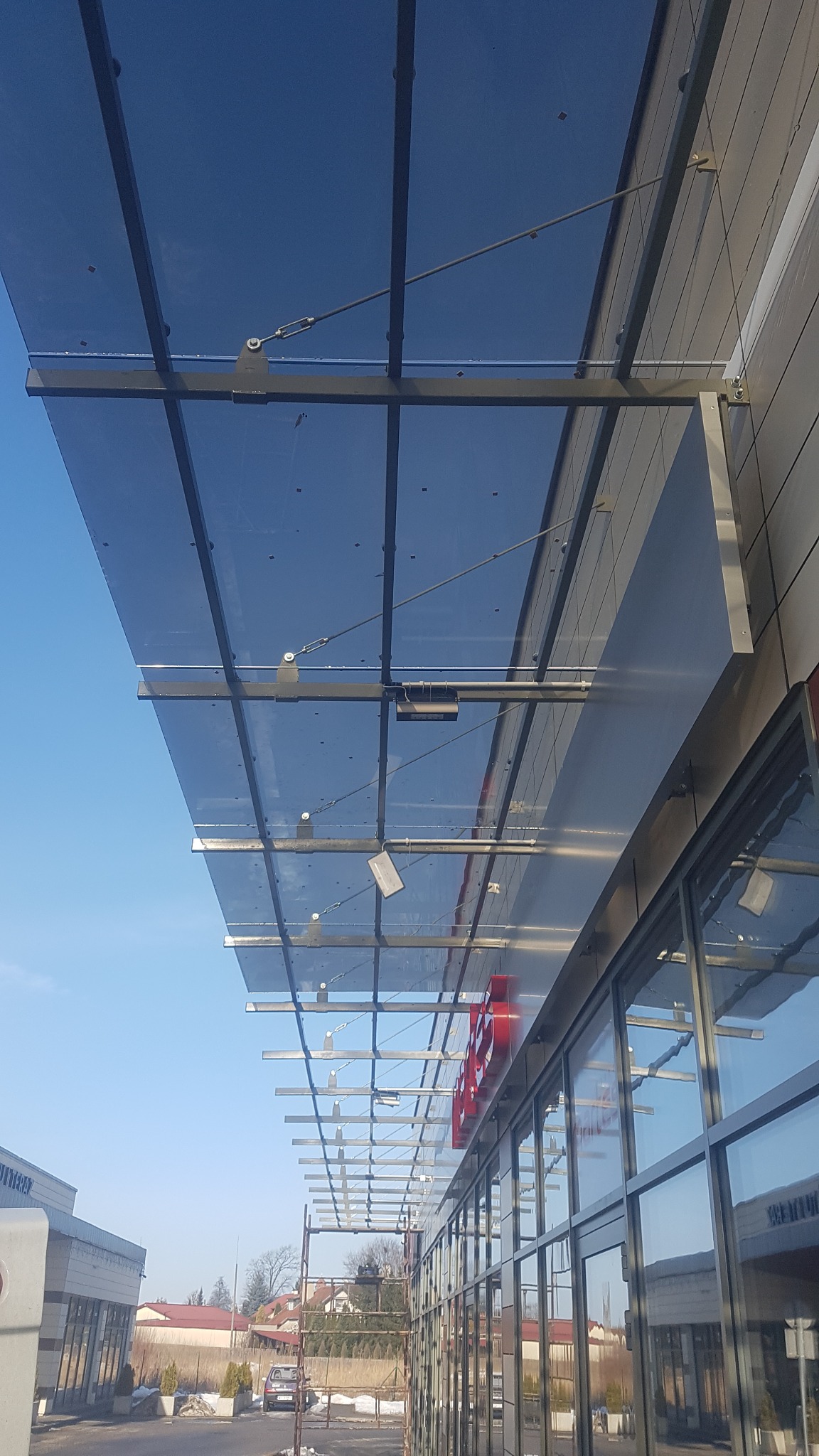 Szklany daszek nad wejściem do budynku, wsparty na metalowej konstrukcji, widok z dołu pod kątem, błękitne niebo w tle.