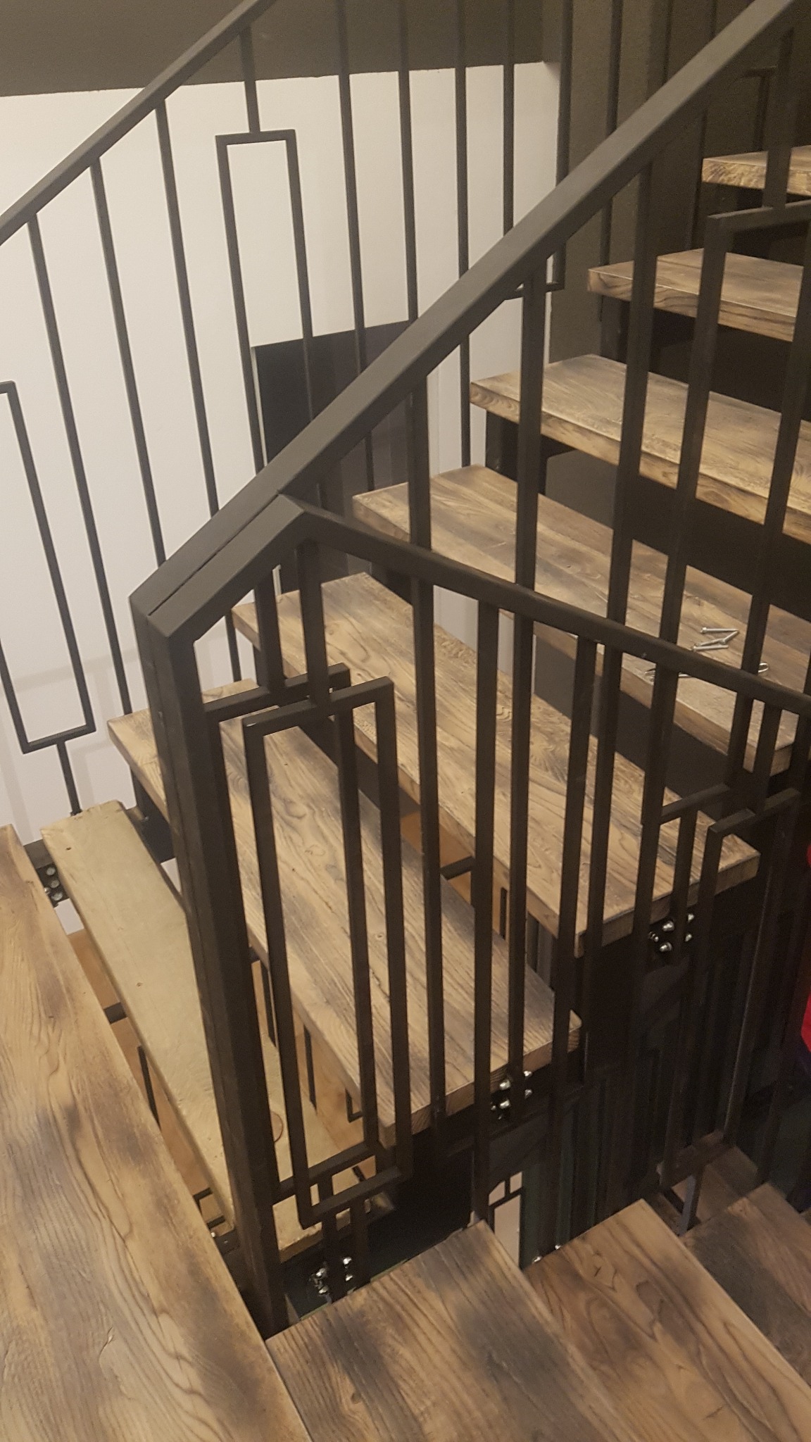Schody z drewnianymi stopniami i czarną metalową balustradą o geometrycznym wzorze, widok z góry na dół.