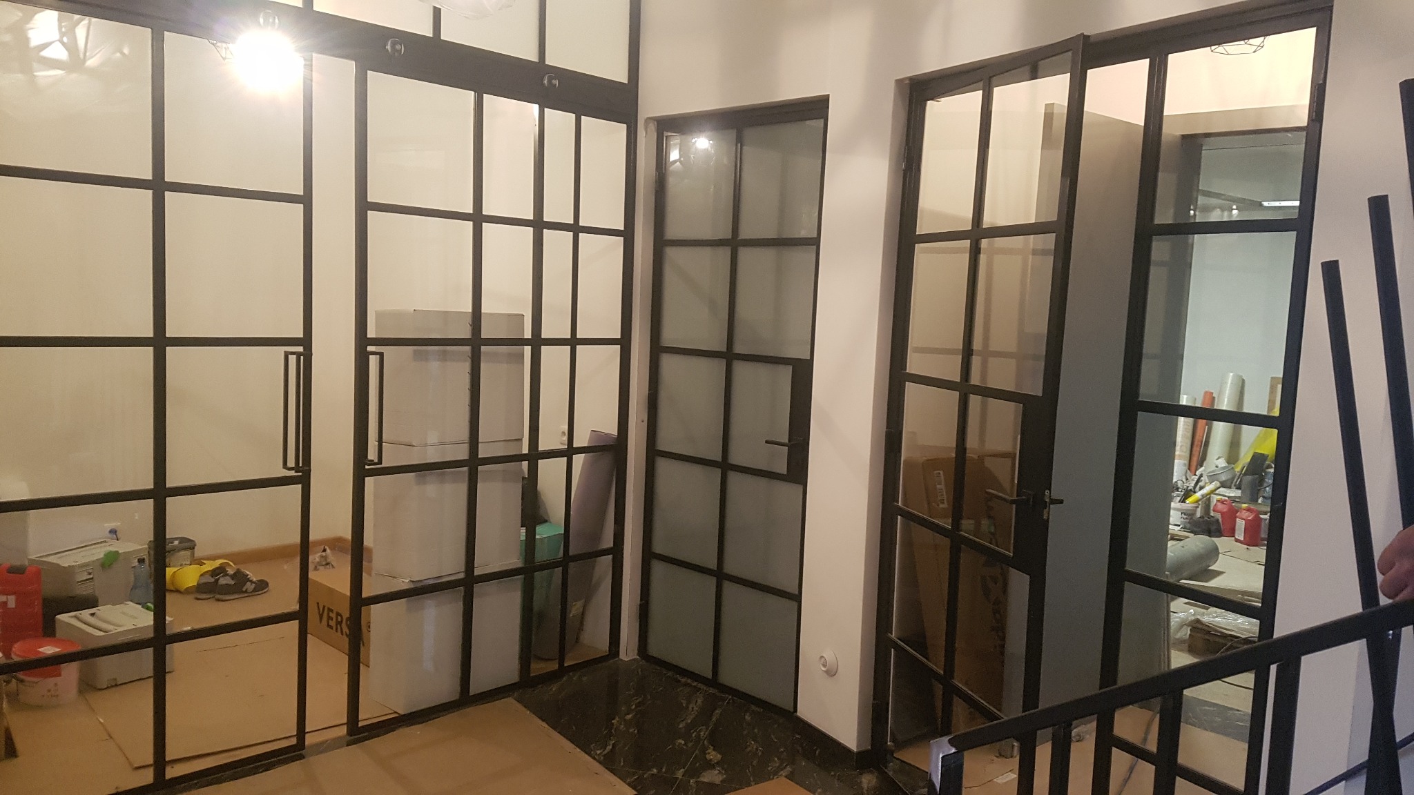 Wnętrze z przeszklonymi ścianami działowymi z czarnymi szprosami, widoczne kartony i narzędzia budowlane sugerujące etap wykończeniowy.