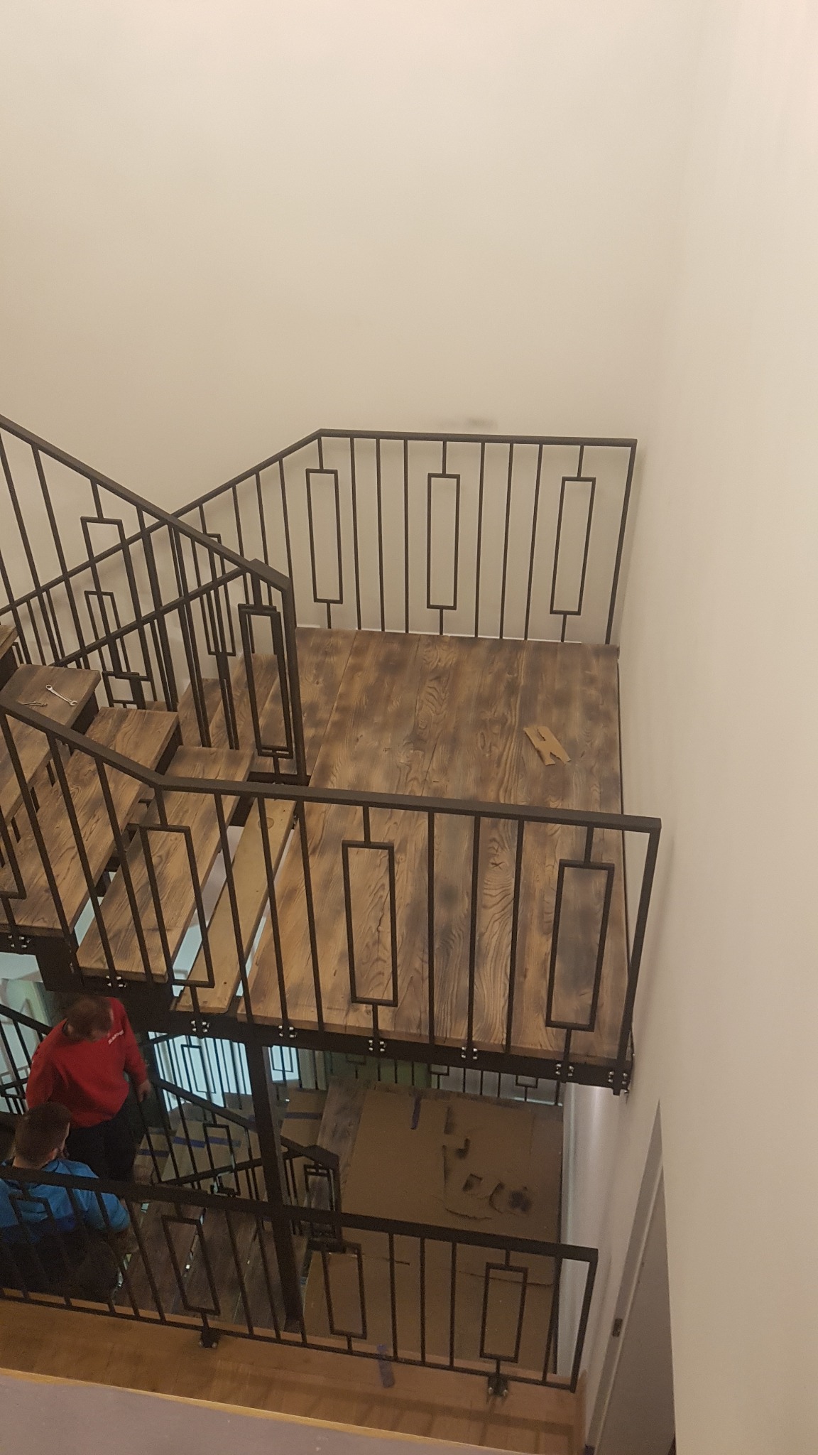 Wnętrze domu z drewnianymi schodami i czarną metalową balustradą o geometrycznym wzorze, widok z góry na schody z osobami wchodzącymi na górę.