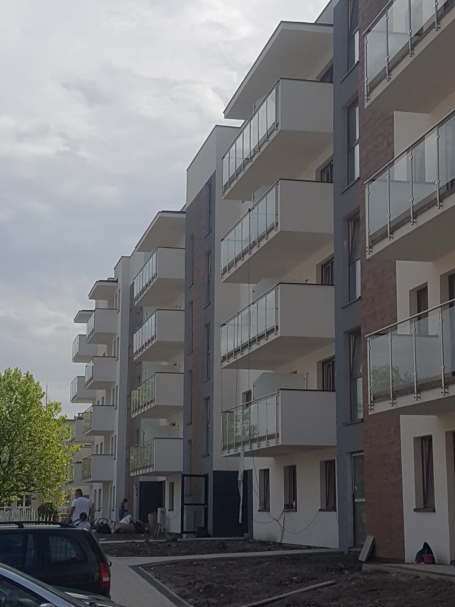 Nowoczesny budynek wielorodzinny z balkonami zabezpieczonymi balustradami szklanymi; widoczni robotnicy na zewnątrz budynku.