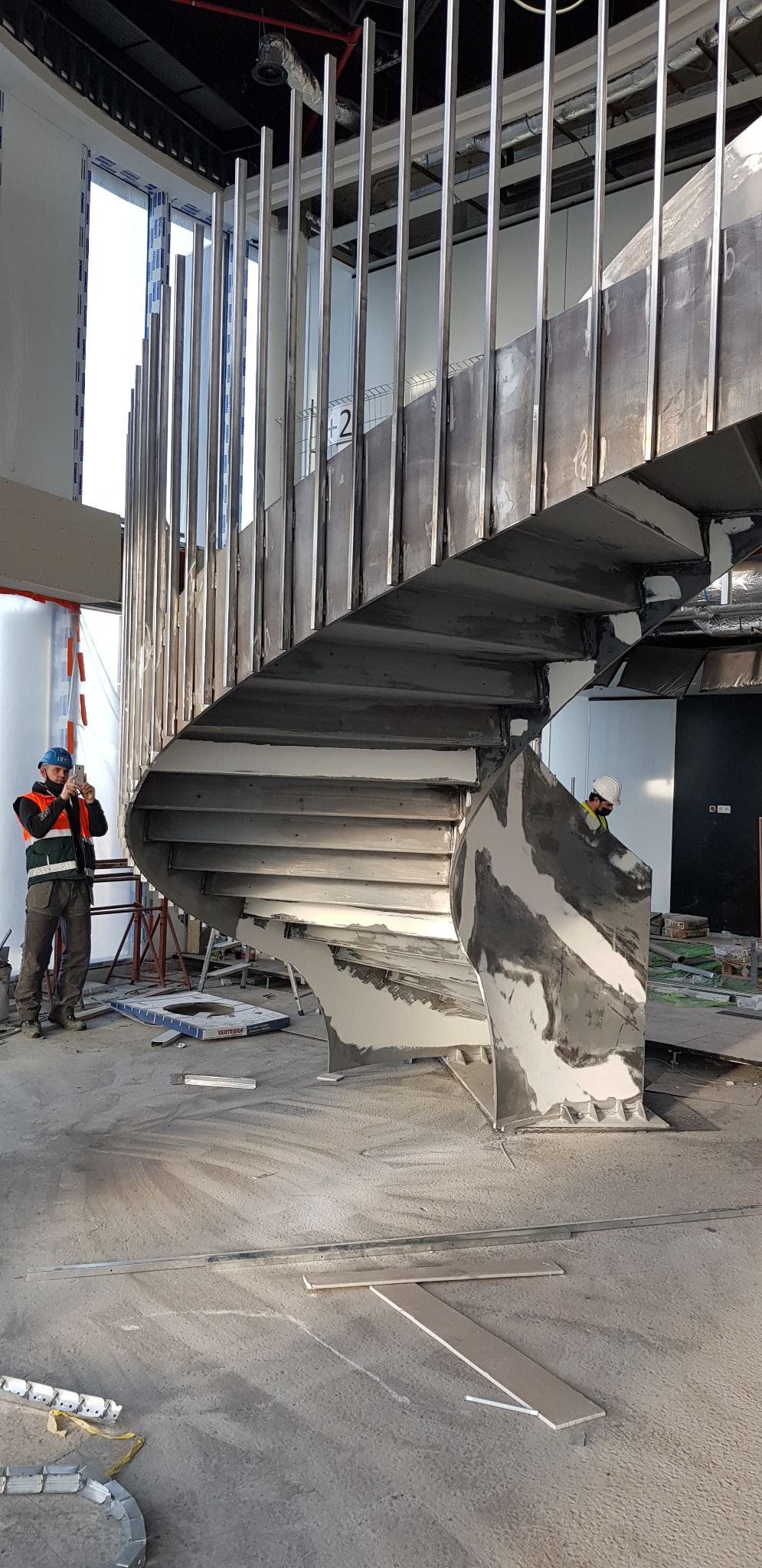 Wewnętrzne schody spiralne z metalu w trakcie montażu, widoczni dwaj pracownicy w kaskach, jeden robiący zdjęcie telefonem, surowy stan wykończenia wnętrza.