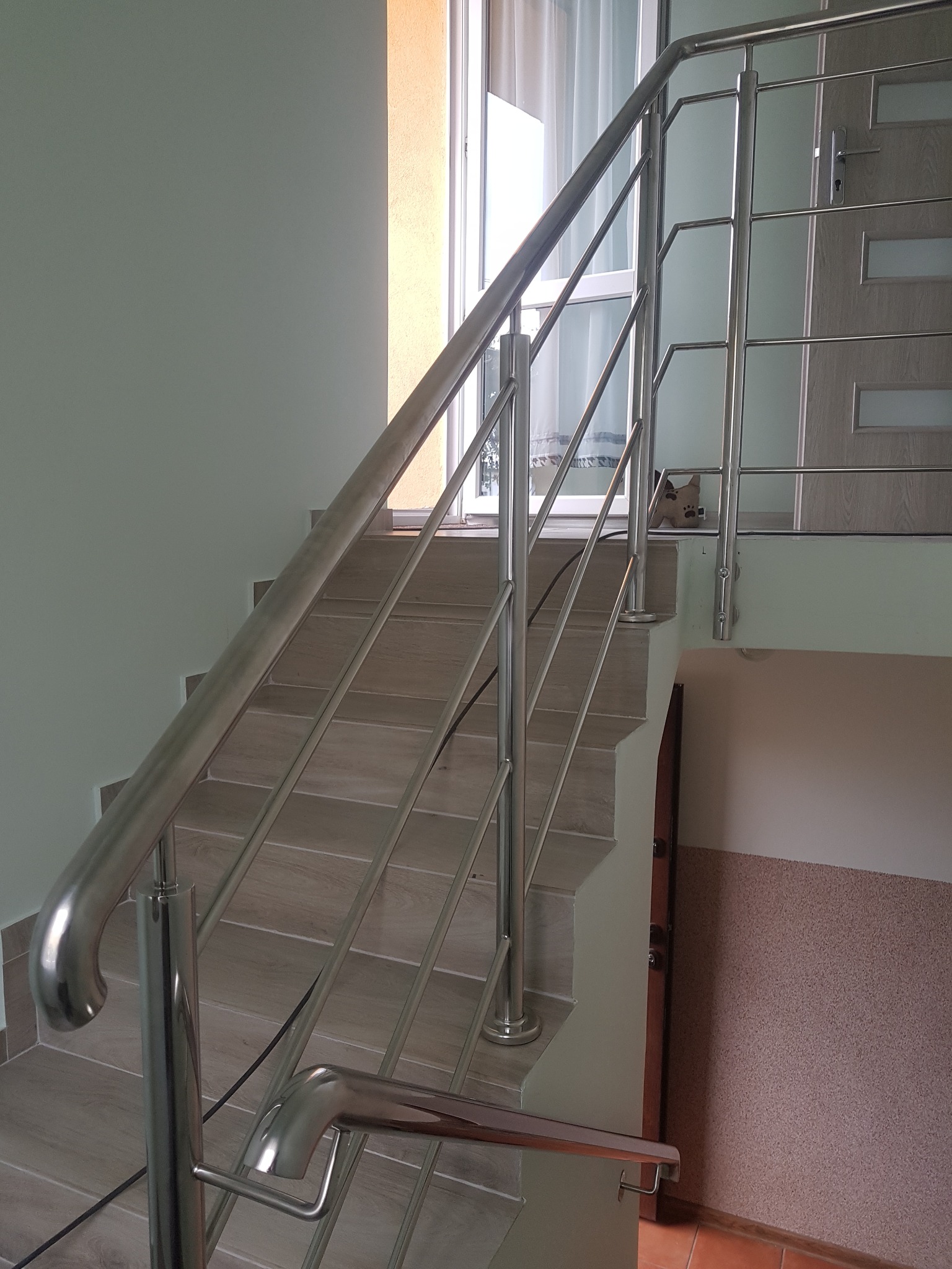 Schody wykończone płytkami w odcieniach beżu w Nidzicy, zabezpieczone nowoczesną balustradą ze stali nierdzewnej, widok z dołu schodów.