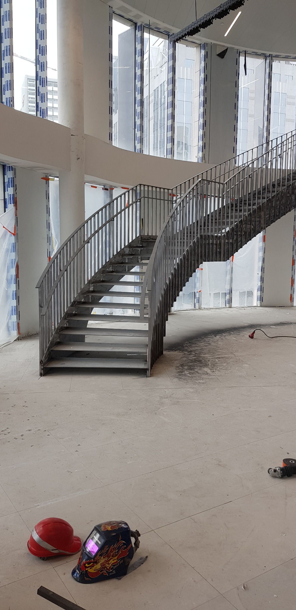 Metalowe schody z szarą balustradą w surowym wnętrzu budynku w trakcie budowy, widoczne narzędzia pracy: kask, maska spawalnicza i szlifierka kątowa.