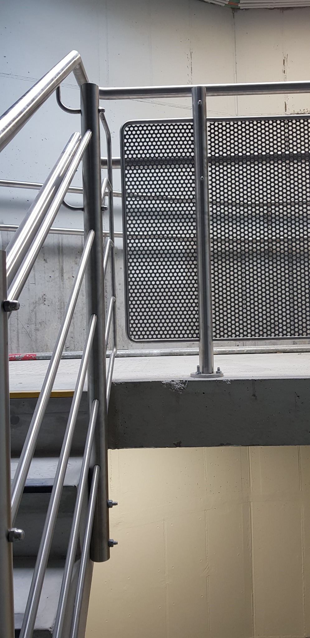 Metalowe schody z balustradą z nierdzewnej stali i perforowanym panelem zabezpieczającym, widoczne mocowania śrubowe.