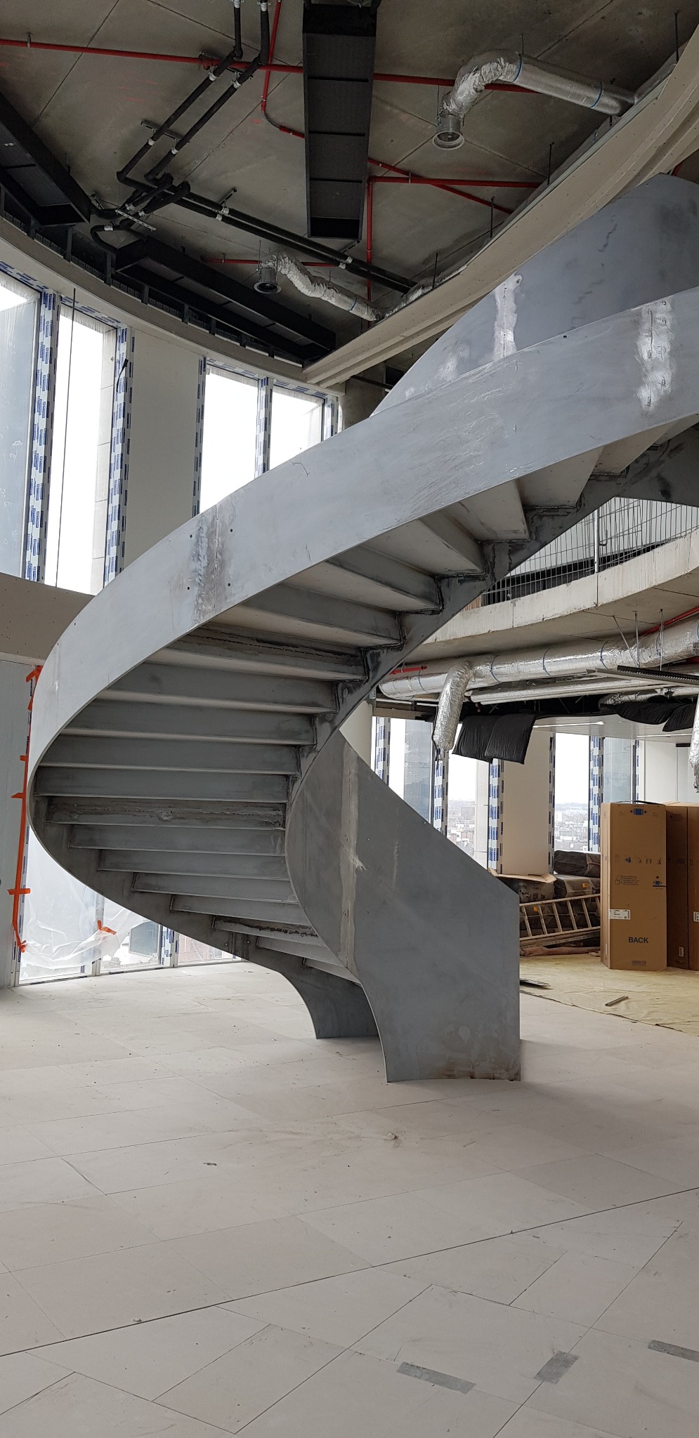 Surowe, metalowe schody spiralne w trakcie montażu w nowoczesnym biurowcu z widokiem na miasto, widoczne instalacje sufitowe i okna.