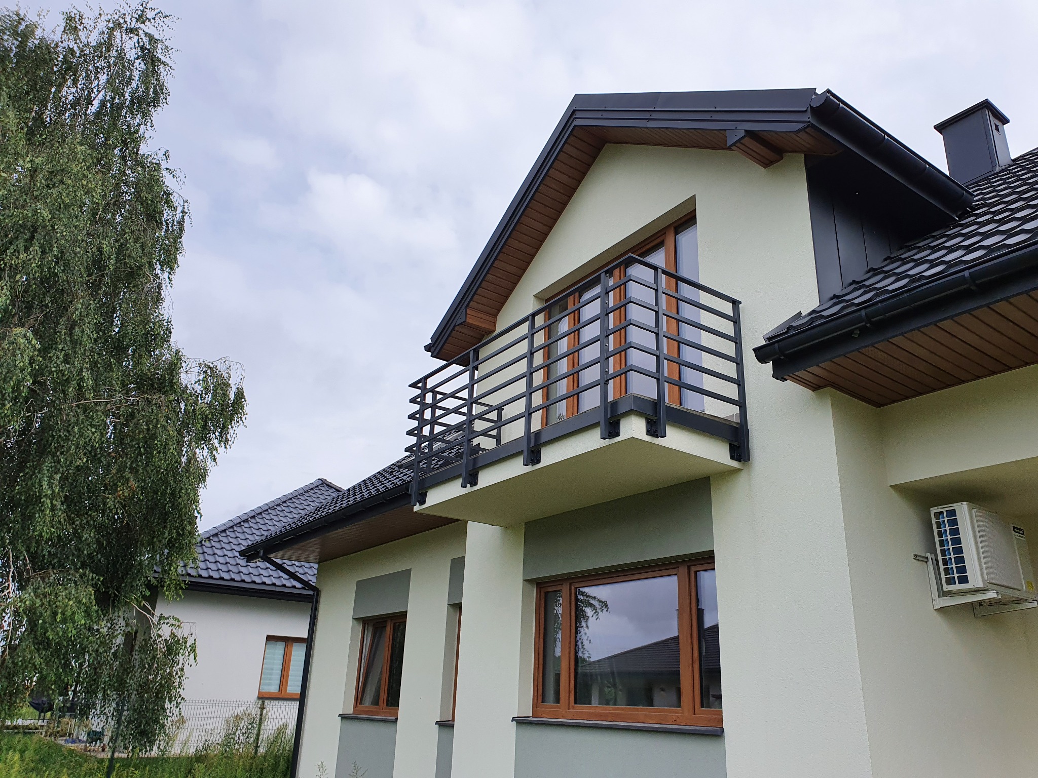 Nowoczesny dom z balkonem zabezpieczonym czarną, metalową balustradą o minimalistycznym designie, widok od dołu na tle pochmurnego nieba.