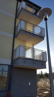 Trzy balkony w nowym budynku z balustradami ze stali nierdzewnej i panelami z mlecznego szkła, widok z dołu na tle błękitnego nieba.