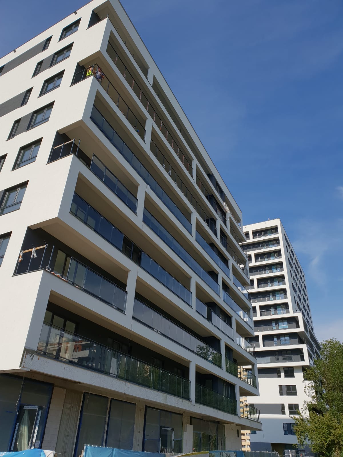 Nowoczesny budynek mieszkalny z białymi balkonami z przeszklonymi balustradami, widok z dołu na tle błękitnego nieba, dwóch pracowników w żółtych kamizelkach na jednym z balkonów.