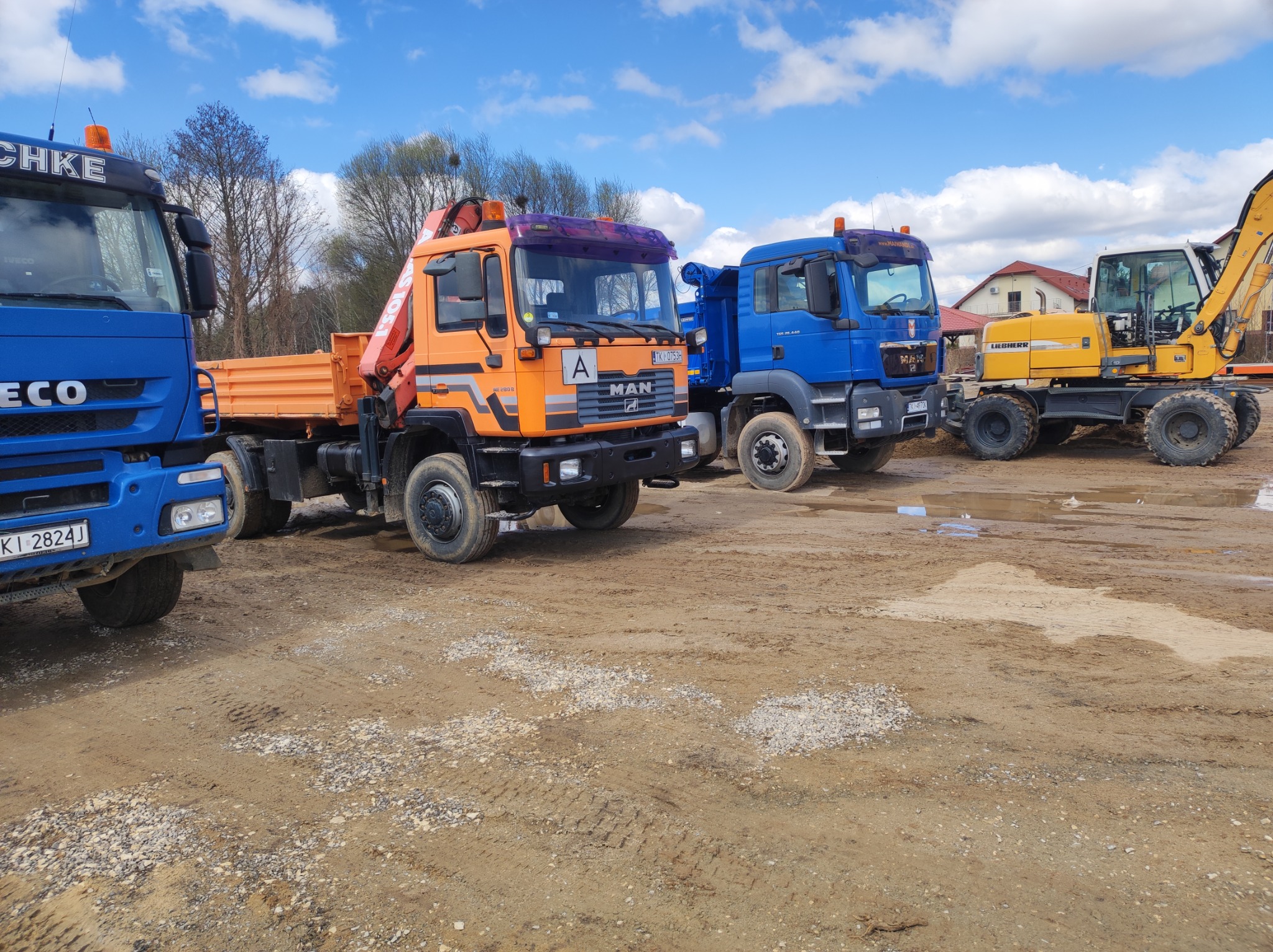 Rząd pojazdów budowlanych na placu: niebieski Iveco, pomarańczowy MAN z dźwigiem oraz żółta koparka kołowa Liebherr, w tle dom jednorodzinny pod błękitnym niebem z chmurami.