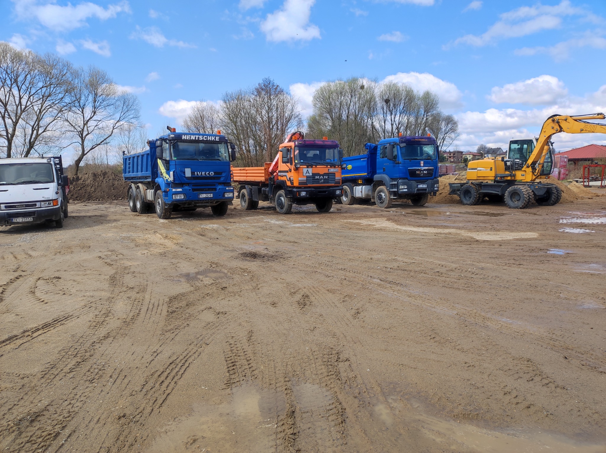 Sprzęt budowlany: trzy ciężarówki różnych marek i kolorów oraz żółta koparka na placu budowy z wyraźnymi śladami opon w błotnistym podłożu, pod błękitnym niebem z chmurami.