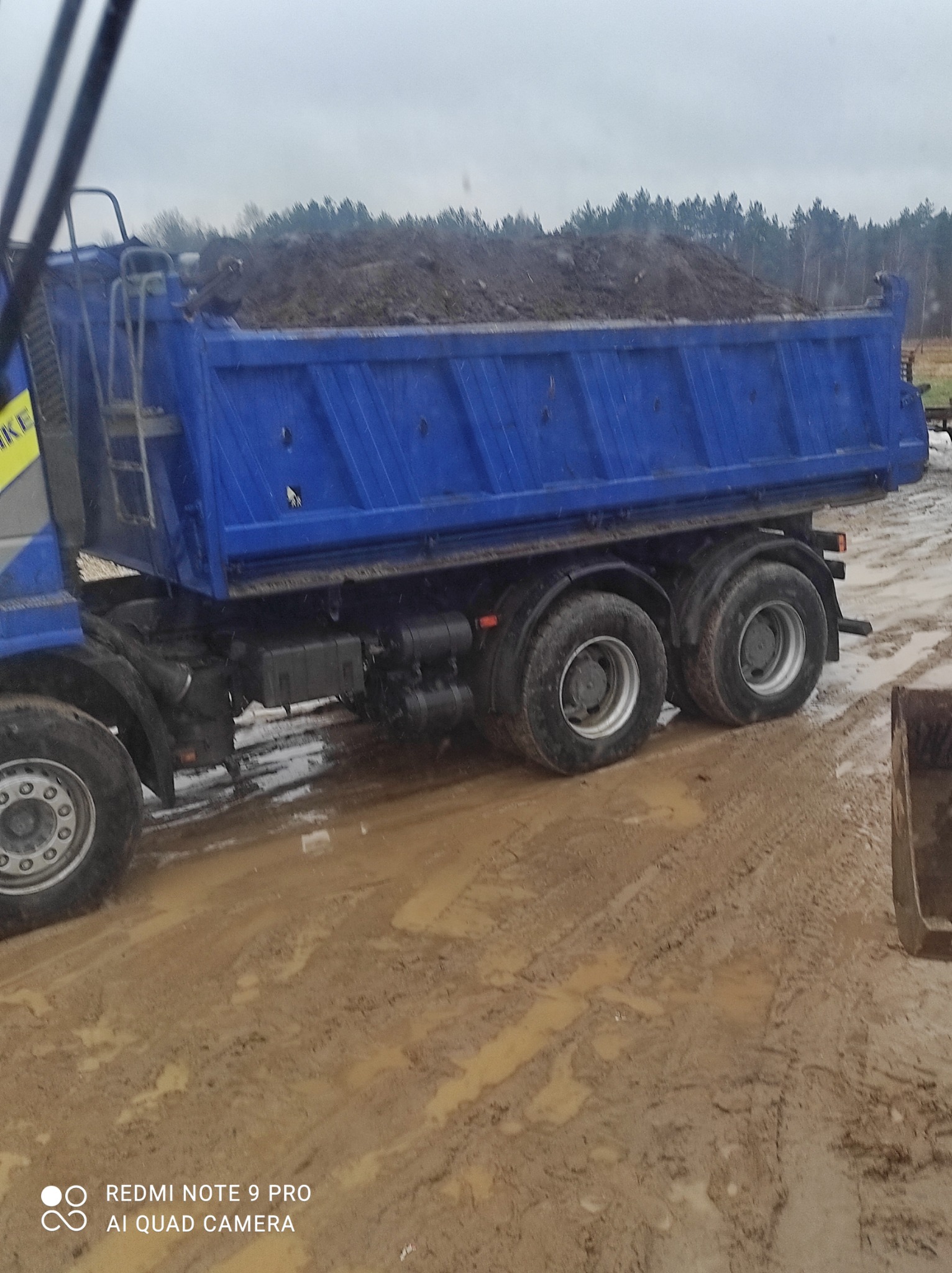 Niebieska wywrotka wypełniona ziemią stoi na błotnistym terenie, w tle widoczne drzewa.