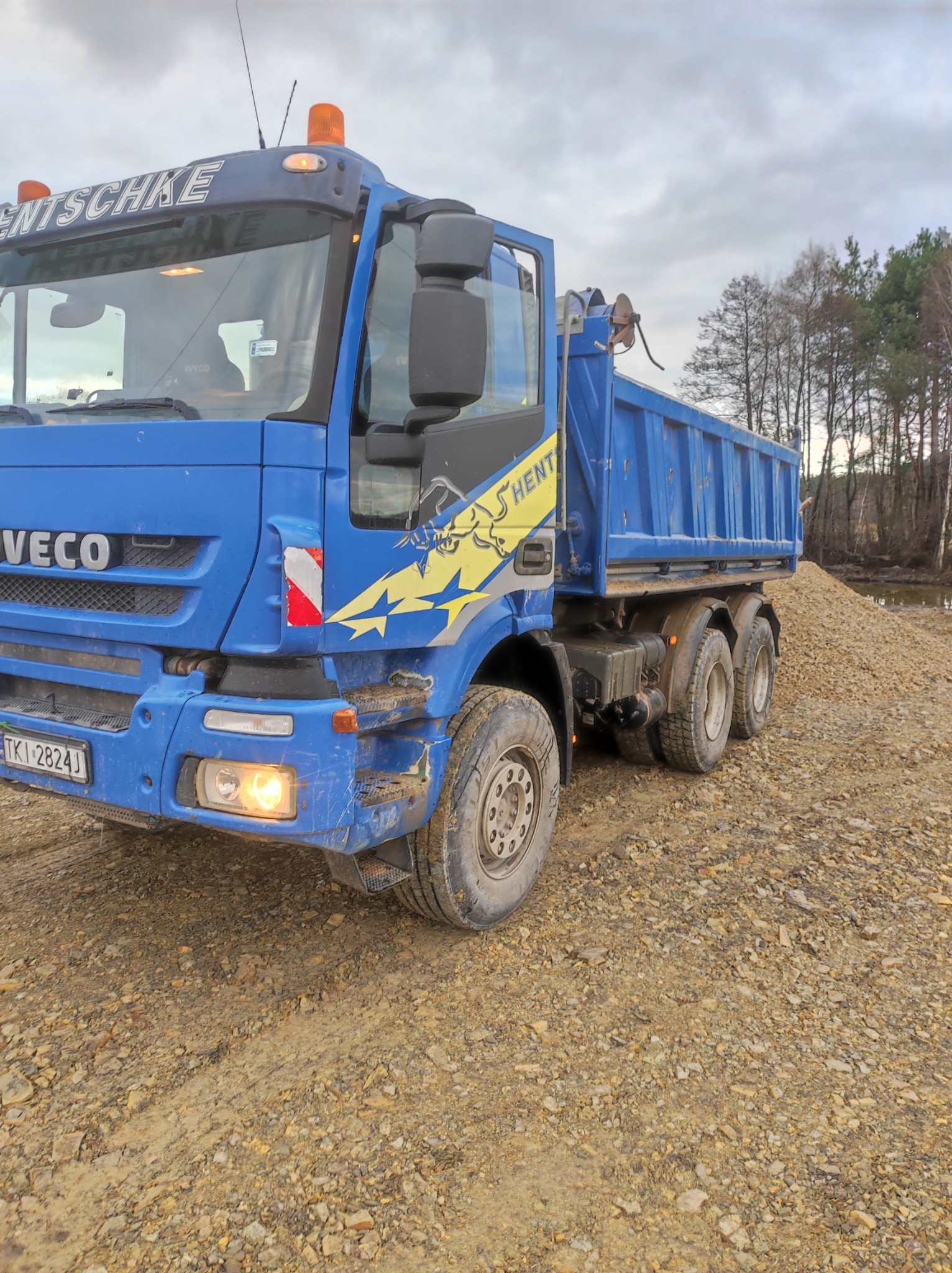 Niebieska wywrotka marki Iveco z żółtymi elementami graficznymi na błotniku, stojąca na gruncie z kamieni i ziemi, z widoczną pryzmą żwiru w tle i drzewami w oddali.