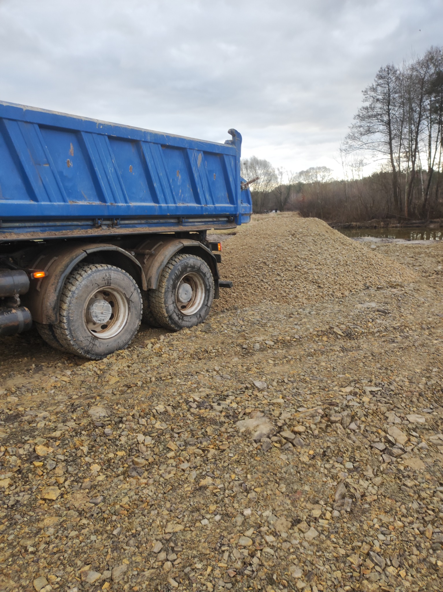 Ciężarówka wywrotka z niebieską skrzynią ładunkową, stojąca obok hałdy żwiru na placu budowy, w tle rzeka i drzewa.