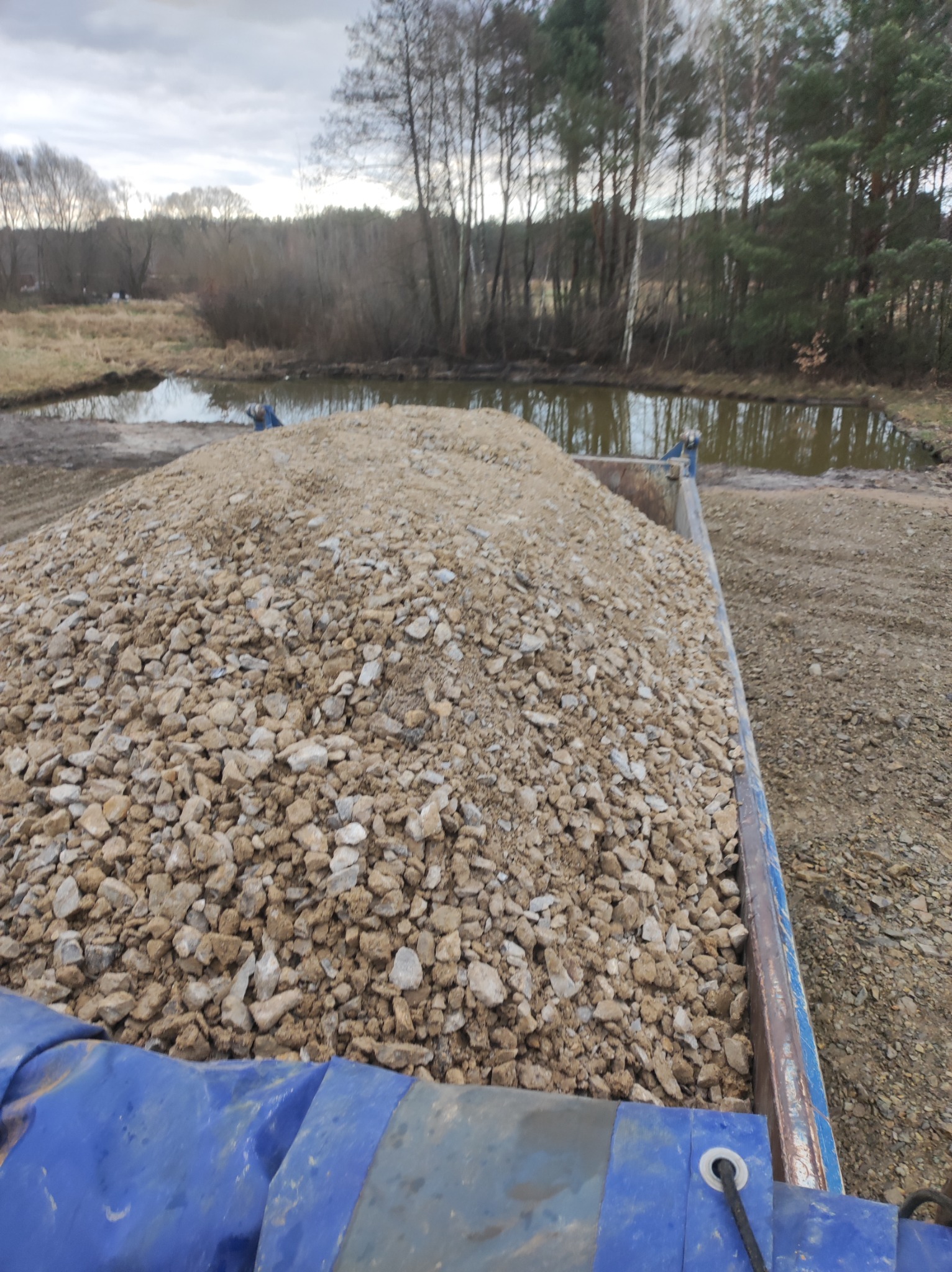 Ładunek kruszywa w skrzyni samochodu ciężarowego, w tle wykopany staw i zalesiony teren w pochmurny dzień.