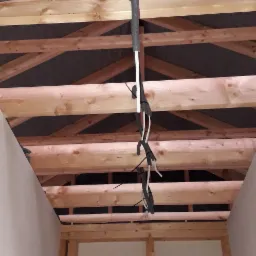 Wiązka przewodów elektrycznych w peszlu przymocowana do drewnianej konstrukcji stropu w surowym stanie, widoczne białe ściany i elementy szkieletu budynku.