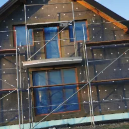 Dom w trakcie prac związanych z izolacją termiczną elewacji, widoczne rusztowania, zabezpieczone okna folią, przyklejone płyty izolacyjne z charakterystycznymi białymi punktami mocowań.