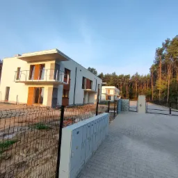 Nowoczesne, dwukondygnacyjne domy jednorodzinne z płaskim dachem, wykończone jasnym tynkiem, z balkonami i drewnianymi elementami elewacji, widziane z poziomu ulicy z chodnikiem z kostki brukowej...