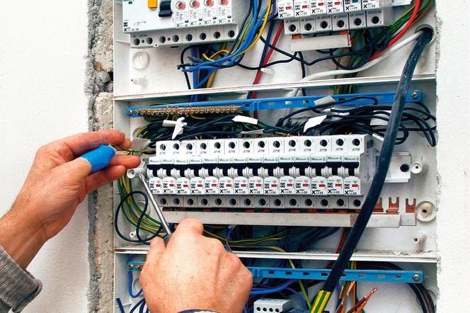 Elektryk dokręca śrubokrętem przewody w skrzynce rozdzielczej z bezpiecznikami, widoczne różnokolorowe kable i automatyczne wyłączniki.