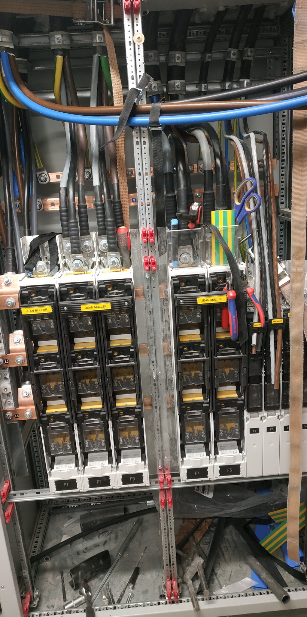Rozdzielnia elektryczna w trakcie montażu, widoczne podłączone przewody, bezpieczniki z etykietami 'JEAN MULLER', narzędzia elektryczne wiszące obok, metalowa konstrukcja szafy i kabelki w różnych...