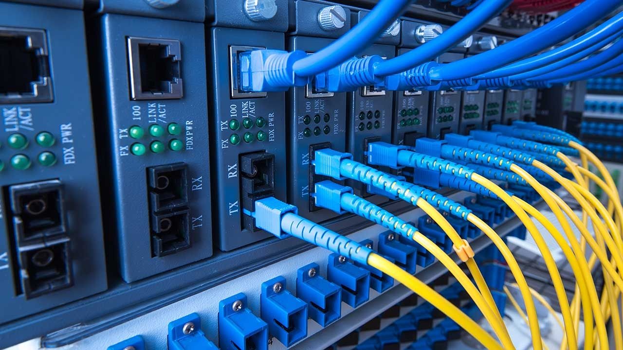 Szereg modułów sieciowych w szafie rack, z podłączonymi niebieskimi i żółtymi kablami światłowodowymi, sygnalizującymi aktywność licznymi zielonymi diodami LED.