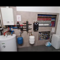 Kompletna kotłownia z piecem gazowym, zbiornikiem CWU, rozdzielaczem ogrzewania podłogowego w zabudowie oraz naczyniami przeponowymi, na tle płytek imitujących kamień.