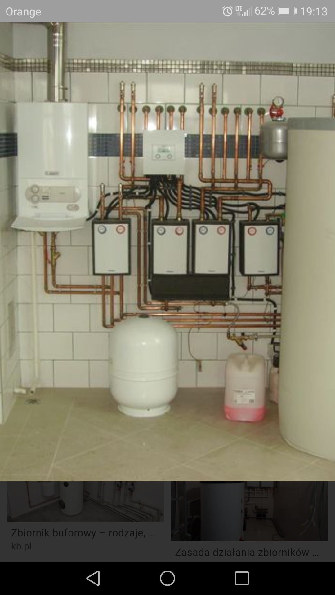 Kotłownia z białym piecem gazowym, rozdzielaczem i zbiornikiem buforowym, instalacja z miedzianych rur, białe płytki na ścianach, kanister z płynem w tle.