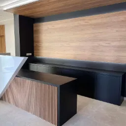 Nowoczesna recepcja z białym, geometrycznym frontem i drewnianymi lamelami, czarne szafki w tle z podświetlanym panelem ściennym z forniru.