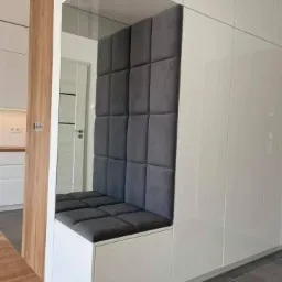 Wnęka z siedziskiem obitym szarą tapicerką w zabudowie szafy w przedpokoju, widoczne lustro i drewniana okładzina ściany.